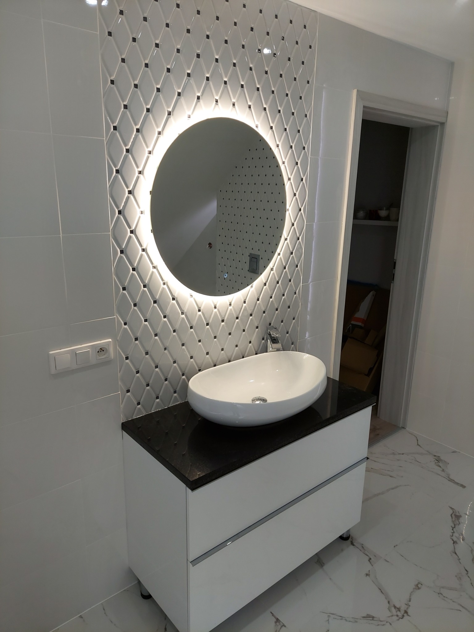Nowoczesna łazienka z owalną umywalką na szafce z czarnym blatem, okrągłe lustro z podświetleniem LED i białe płytki z czarnymi akcentami na ścianie.