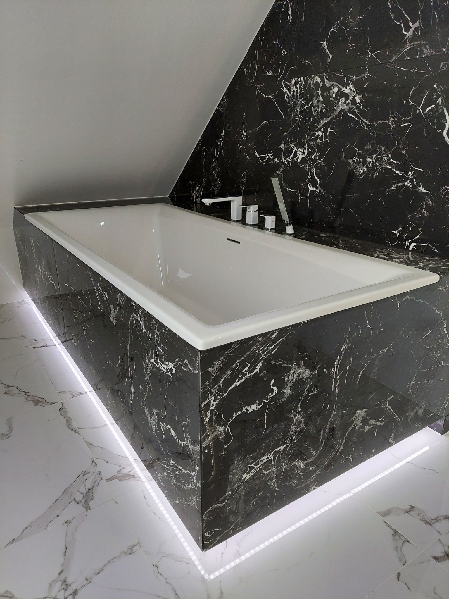 Nowoczesna wanna obudowana czarnym marmurem z białymi żyłami, podświetlona listwą LED u dołu, na tle białej marmurowej podłogi i ściany z czarnego marmuru.