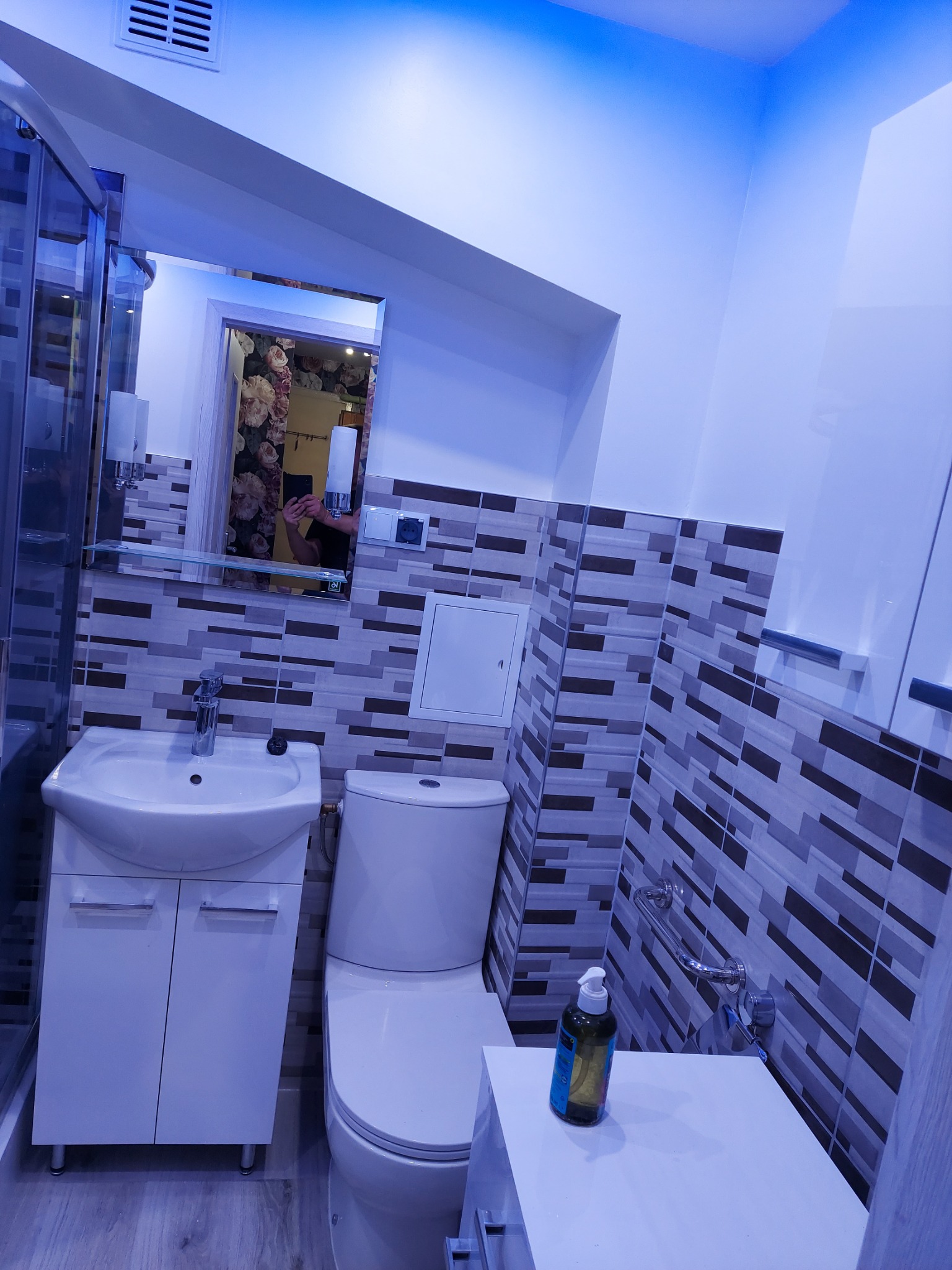 Wykończona łazienka w odcieniach bieli i szarości z akcentami niebieskiego oświetlenia LED, widoczna umywalka z szafką, toaleta oraz fragment kabiny prysznicowej. Na blacie stoi butelka z dozownikiem.