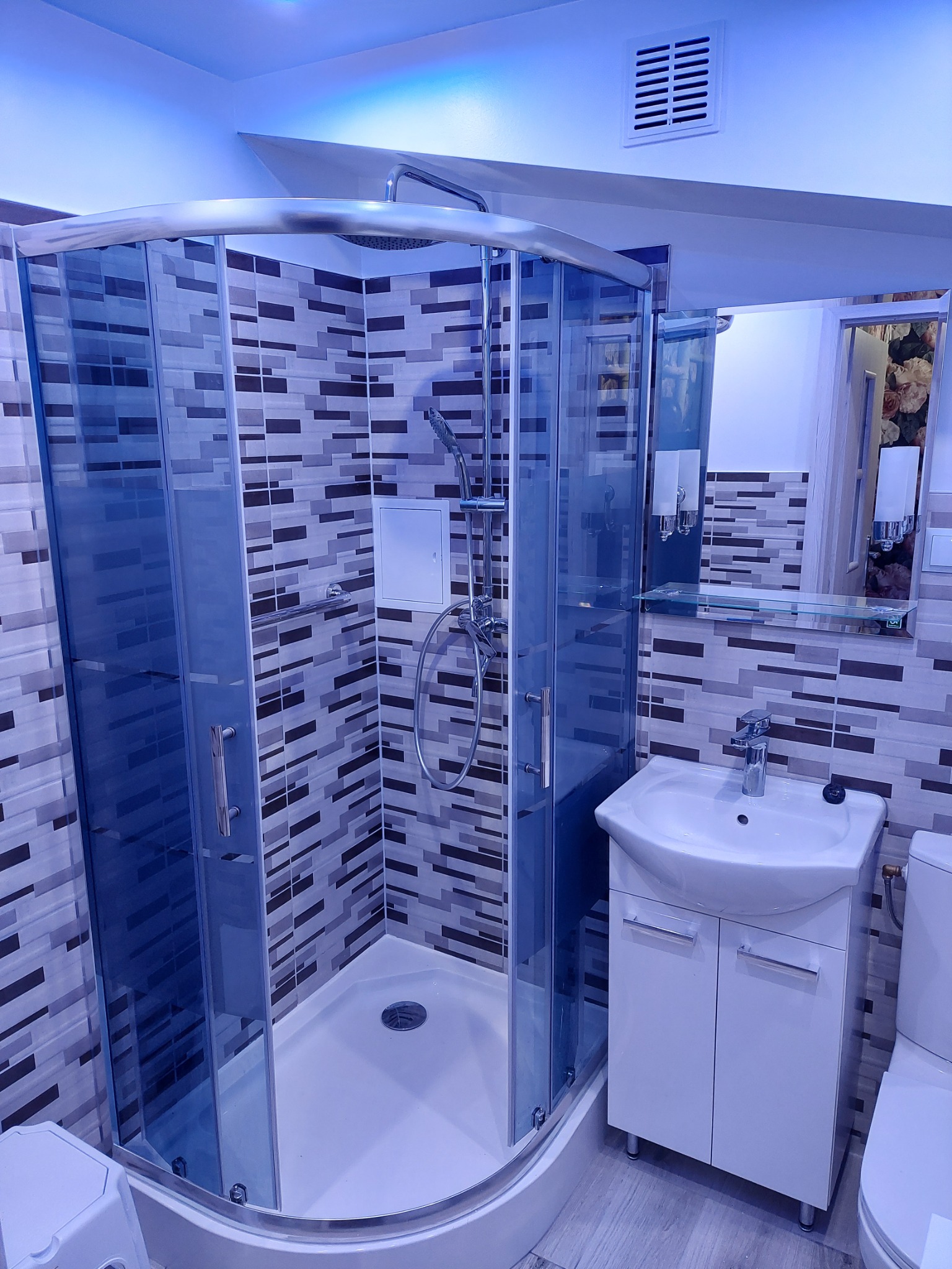Nowoczesna kabina prysznicowa z chromowanymi elementami, umywalka z szafką i toaleta w łazience wykończonej mozaiką w odcieniach szarości i bieli, oświetlenie sufitowe w chłodnej tonacji.