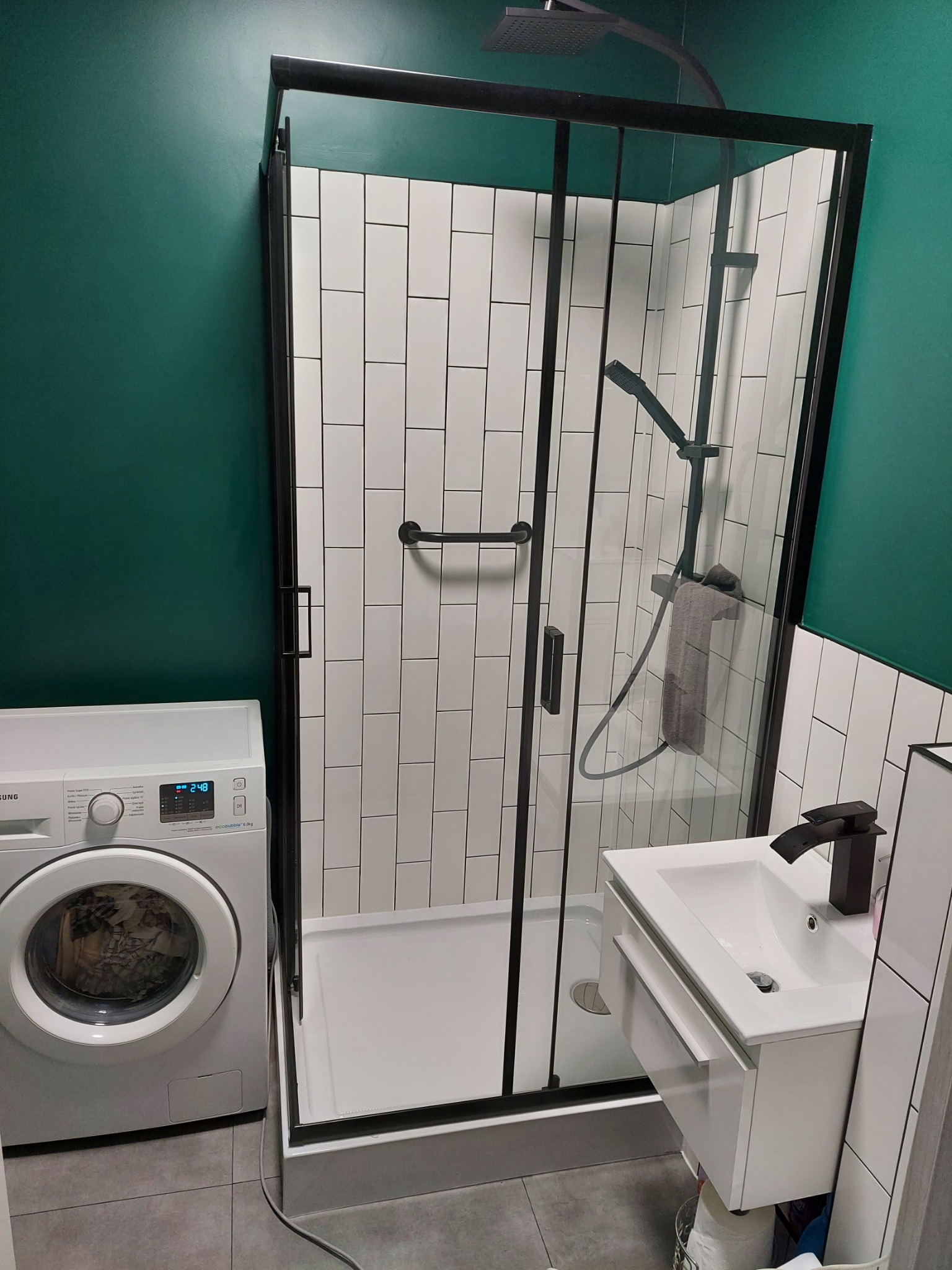 Nowoczesna łazienka z kabiną prysznicową typu walk-in z czarnymi profilami, umywalką z szafką i pralką Samsung, ściany wyłożone białymi, podłużnymi płytkami oraz pomalowane na ciemny zielony kolor.