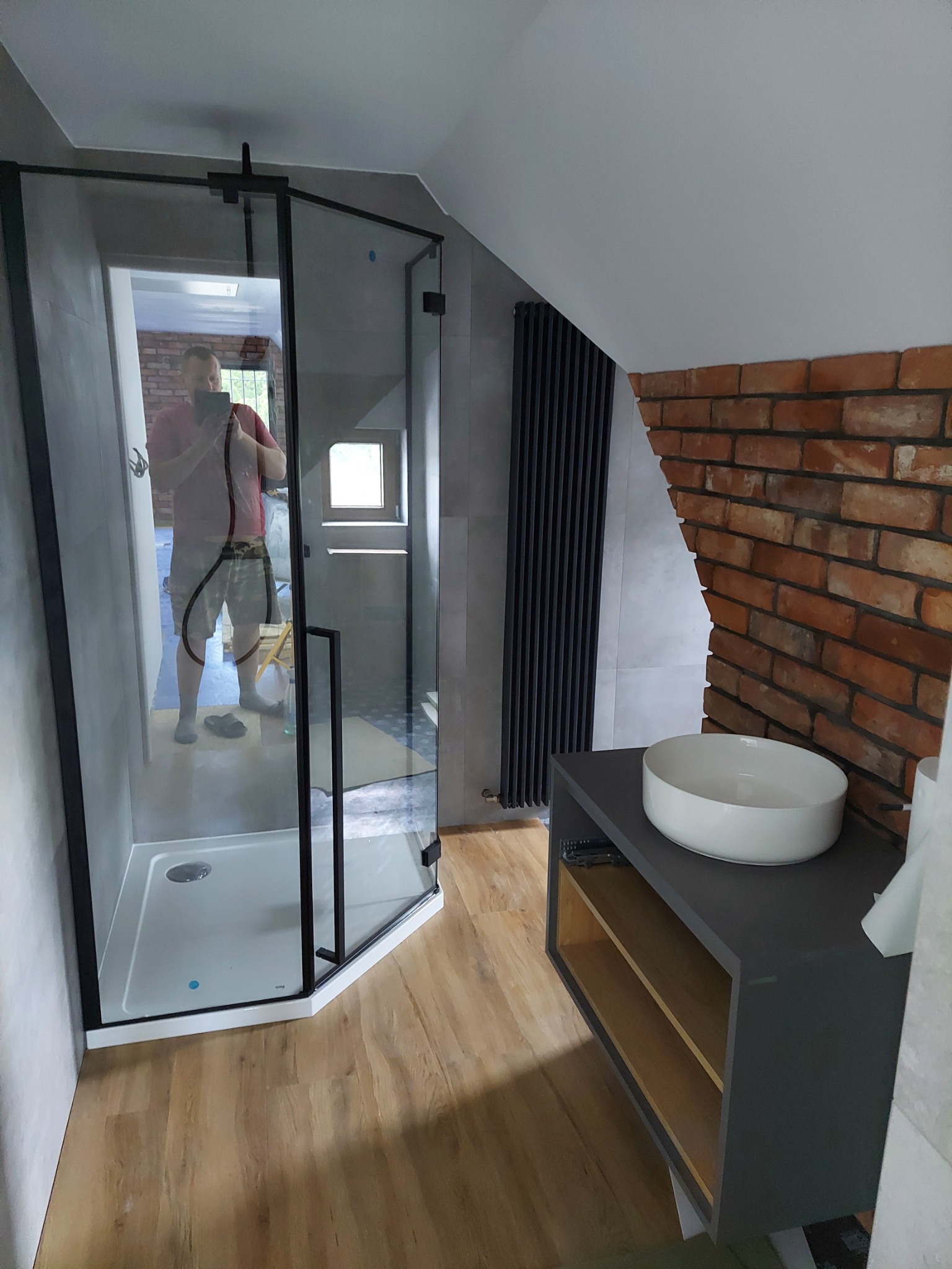 Nowoczesna łazienka na poddaszu z kabiną prysznicową z czarnymi profilami, umywalką nablatową na szarej szafce i ścianą z cegły; odbicie fotografa w szkle kabiny.