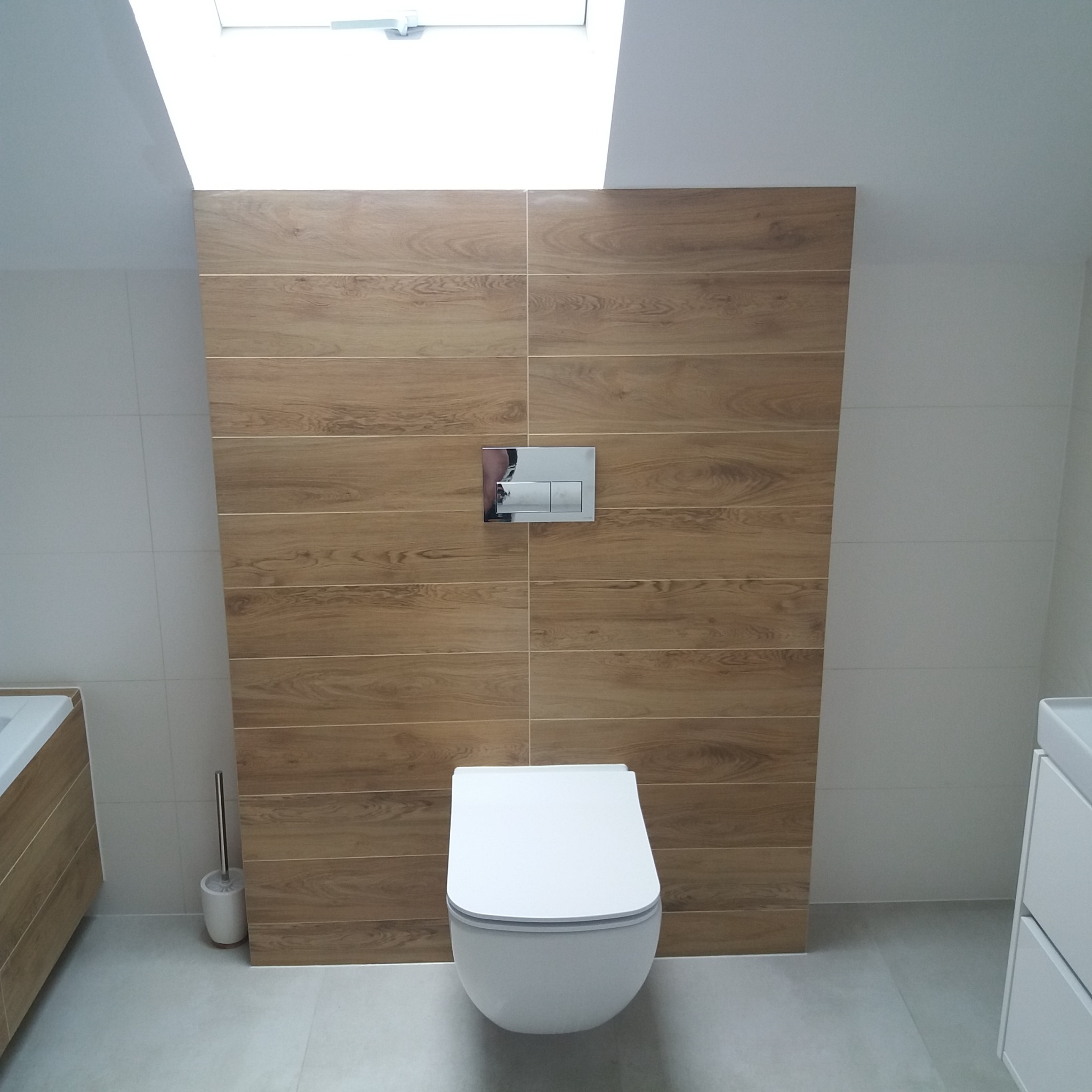 Nowoczesna łazienka z wiszącą toaletą na tle ściany wyłożonej płytkami imitującymi drewno, z chromowanym przyciskiem spłukującym, widok na jasne wnętrze z oknem dachowym.