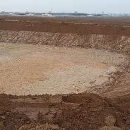 Szeroki wykop wyłożony geokratą z widocznymi śladami erozji, przygotowany pod zbiornik retencyjny na tle zabudowań gospodarczych w pochmurny dzień.