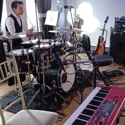 Cinnamon Live Cover Band - Perkusista w trakcie przygotowań do występu, widoczny zestaw perkusyjny, czerwony keyboard Nord, gitara oraz oświetlenie sceniczne.