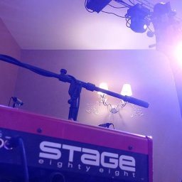 Cinnamon Live Cover Band - Czerwony keyboard Stage Eighty Eight z mikrofonem na statywie, oświetlony reflektorami, w tle ściana z dekoracyjnym kinkietem i fragment portretu kobiety.