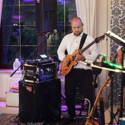 Cinnamon Live Cover Band - Gitarzysta w białej koszuli gra na elektrycznej gitarze na scenie podczas występu, widoczny sprzęt nagłośnieniowy i oświetlenie sceniczne, w tle okno z białymi zasłonami i wzorzysta tapeta.