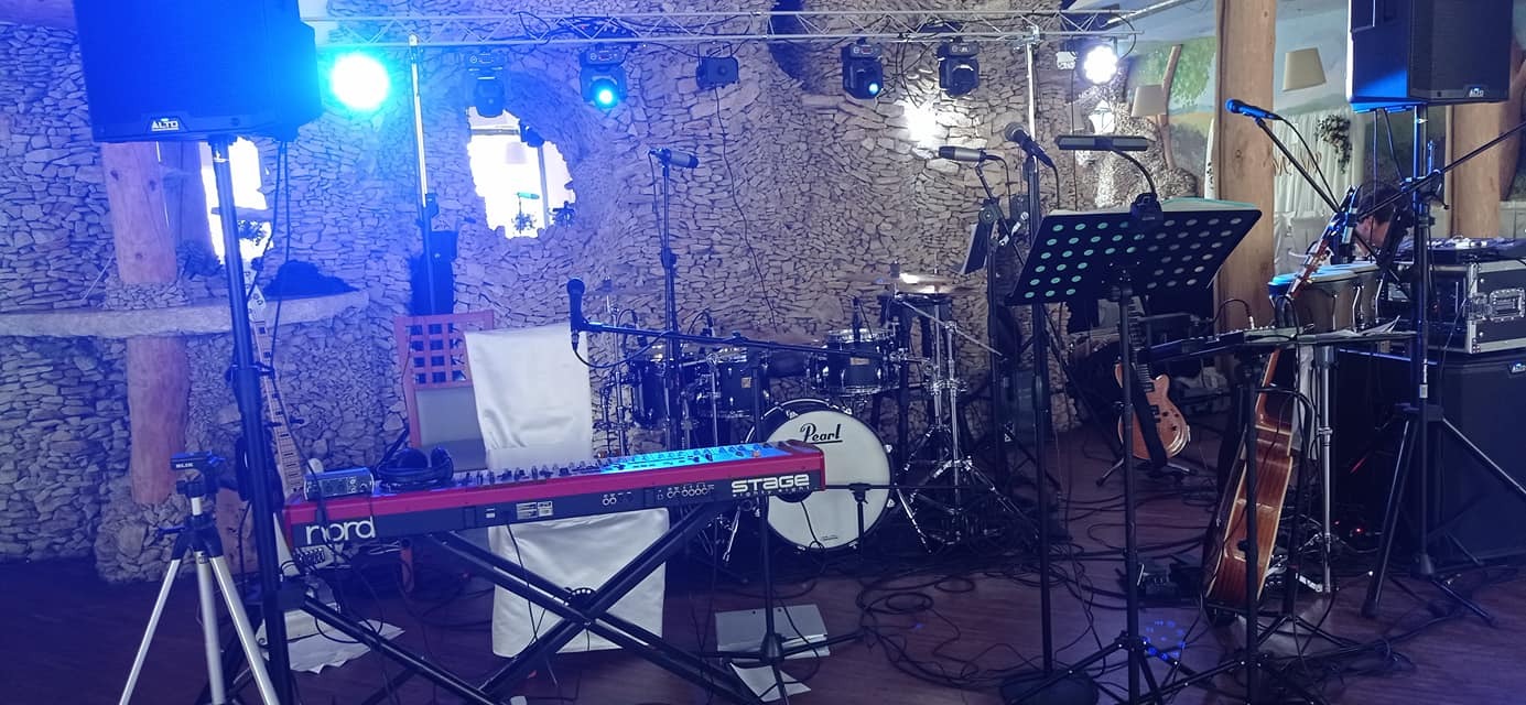Przygotowany do występu zestaw instrumentów muzycznych na scenie z kamiennym tłem, oświetlony niebieskim światłem, widoczne keyboard Nord, perkusja Pearl Stage, gitary i mikrofony.