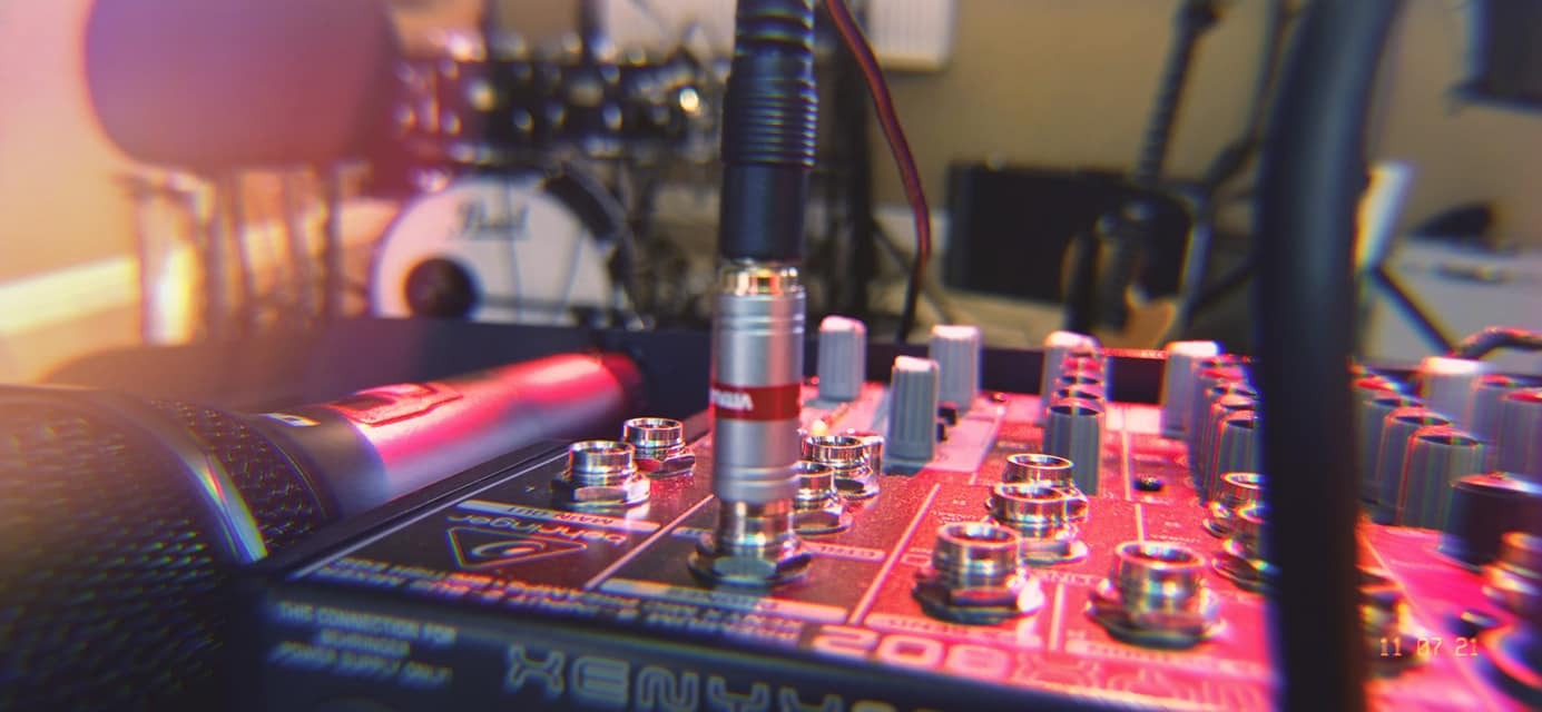 Zbliżenie na mikser audio Behringer XENYX 202, z wpiętym kablem XLR i widocznym mikrofonem oraz perkusją w tle, oświetlone czerwonym światłem.