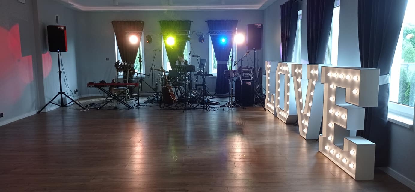 Sala weselna przygotowana na przyjęcie z podświetlanymi literami LOVE, rozstawionym sprzętem muzycznym: perkusja, gitara, keyboard, kolumny nagłośnieniowe, oświetlenie sceniczne.