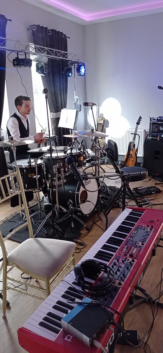 Perkusista w trakcie przygotowań do występu, widoczny zestaw perkusyjny, czerwony keyboard Nord, gitara oraz oświetlenie sceniczne.