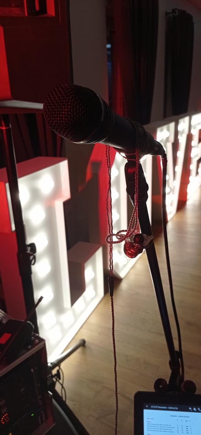 Mikrofon na statywie z wiszącymi słuchawkami dousznymi, w tle podświetlane litery układające się w słowo 'LIVE', sprzęt audio na podłodze.