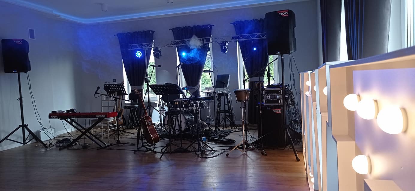 Scena przygotowana do występu: perkusja, gitara, keyboard, nagłośnienie i oświetlenie sceniczne ustawione w sali z dekoracyjnymi zasłonami i oświetleniem.