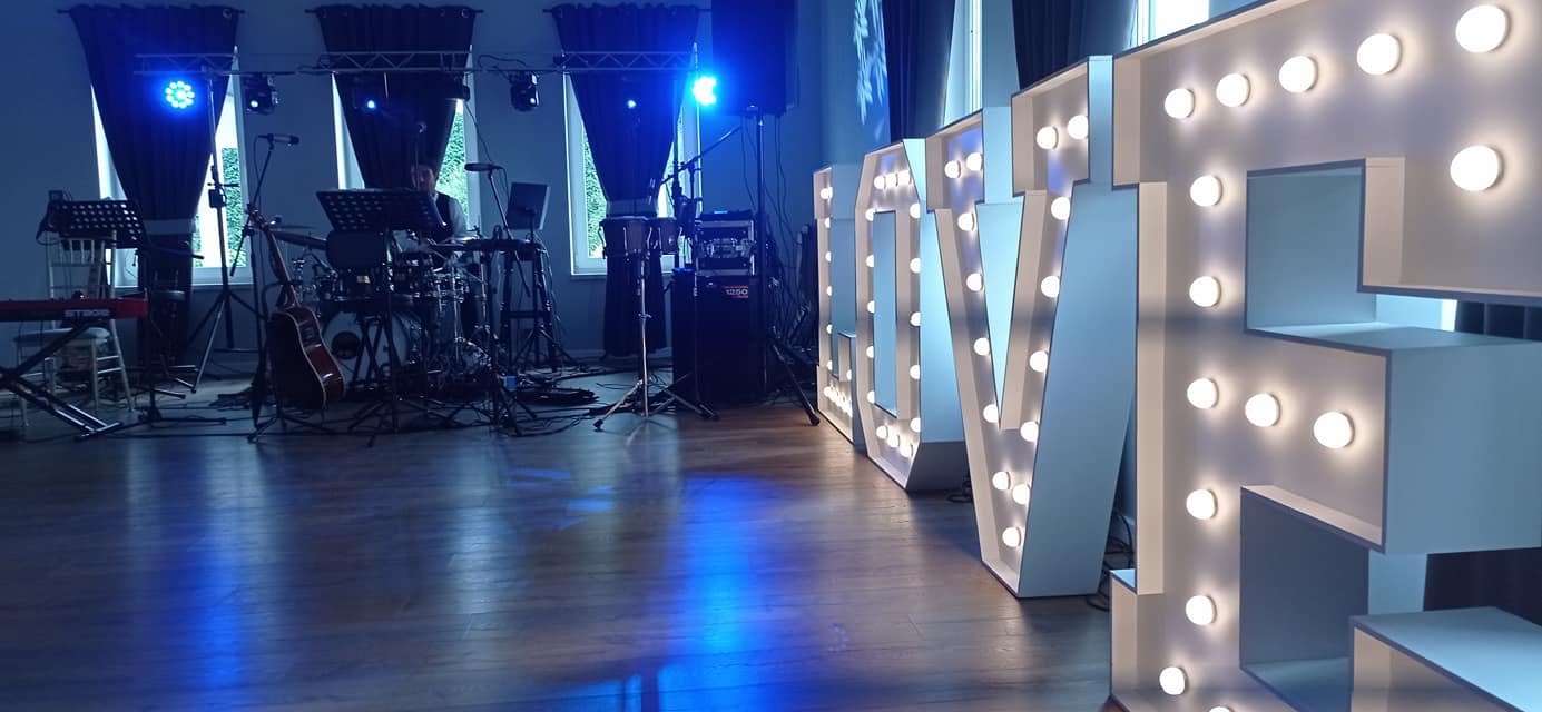 Scena z instrumentami muzycznymi: perkusja, keyboard, gitara akustyczna, oświetlenie sceniczne i dekoracyjne litery podświetlane żarówkami, ustawione w sali z oknami.