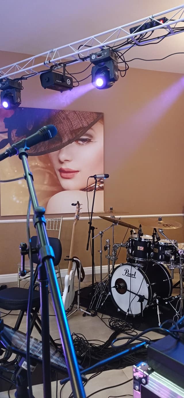 Scena z instrumentami muzycznymi: perkusja Pearl, gitara basowa, mikrofony. W tle oświetlenie sceniczne i plakat z portretem kobiety w kapeluszu.