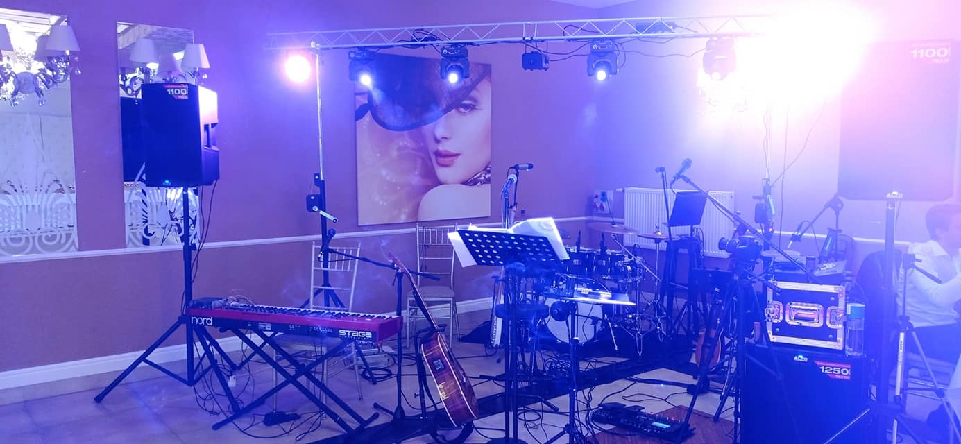 Sprzęt muzyczny zespołu weselnego: keyboard Nord Stage, gitara akustyczna, perkusja oraz nagłośnienie na tle plakatu z wizerunkiem kobiety.