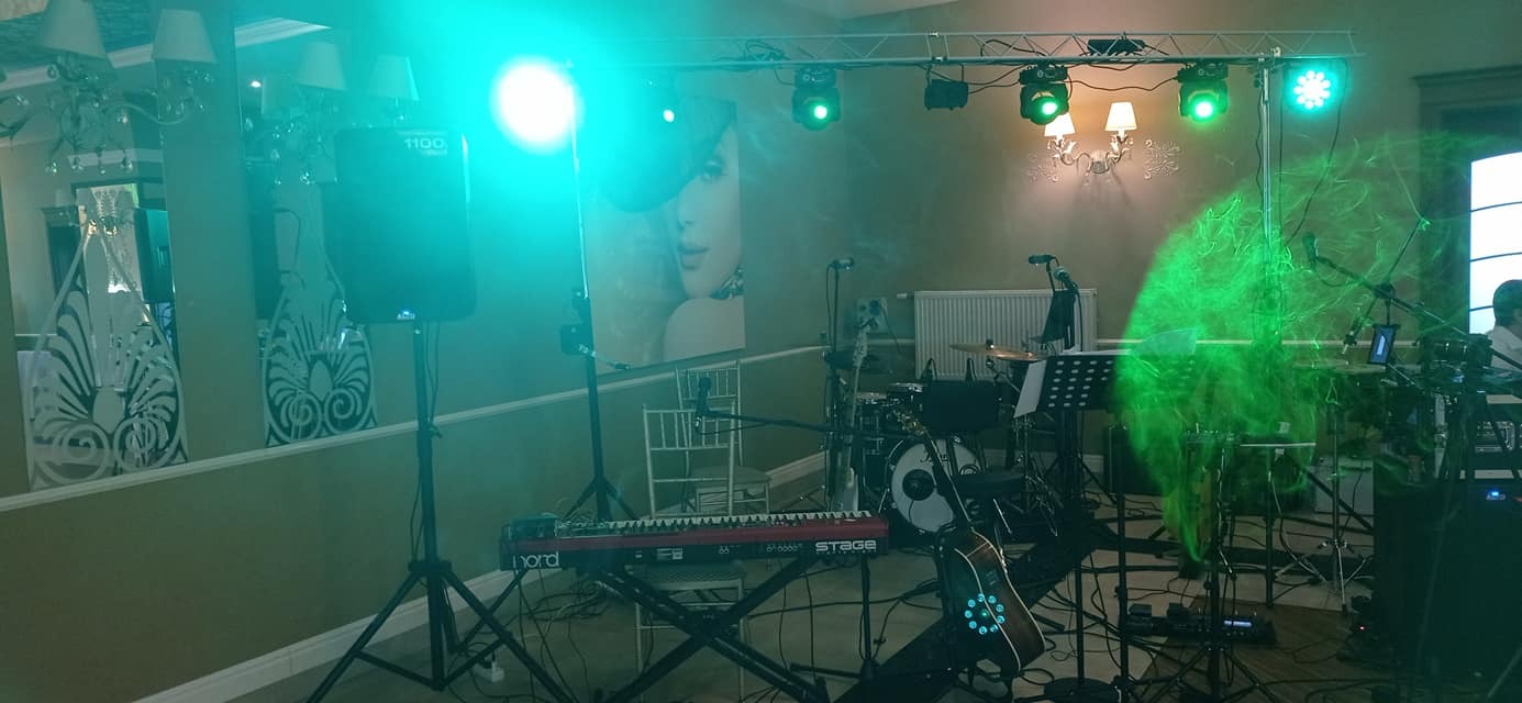 Scena z instrumentami muzycznymi, w tym keyboard Nord Stage, perkusja i gitara akustyczna z podświetleniem, ustawiona w sali z dekoracyjnym oświetleniem i plakatem kobiety w tle, oświetlona...