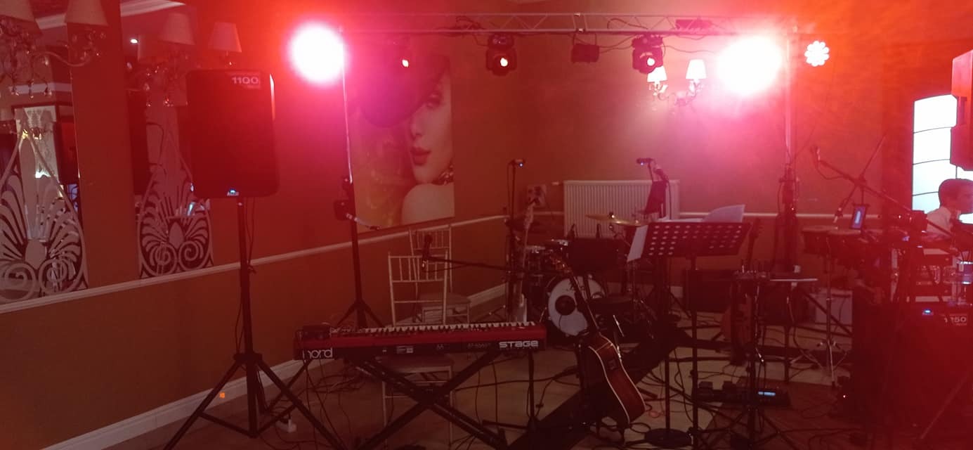 Scena z instrumentami muzycznymi i sprzętem nagłośnieniowym, przygotowana do występu, oświetlona czerwonym światłem, widoczny keyboard Nord Stage, perkusja i kolumny głośnikowe.
