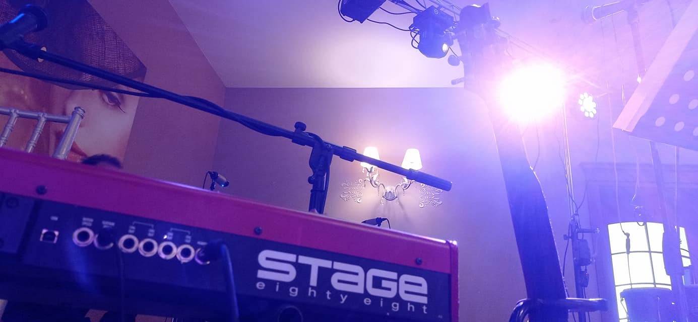 Czerwony keyboard Stage Eighty Eight z mikrofonem na statywie, oświetlony reflektorami, w tle ściana z dekoracyjnym kinkietem i fragment portretu kobiety.