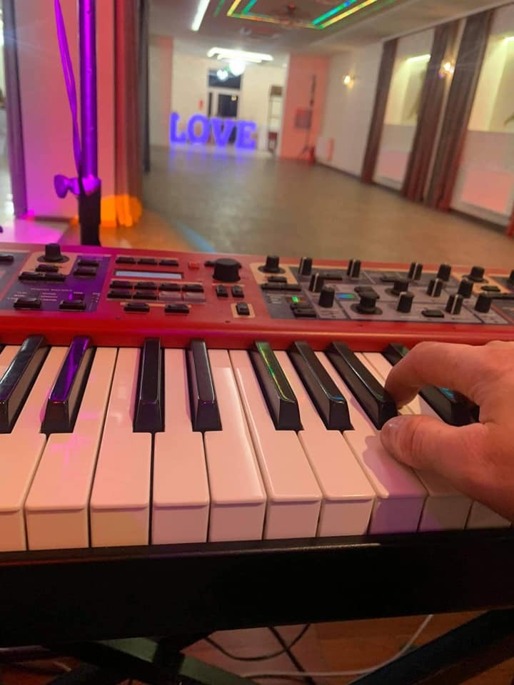 Dłoń grająca na czerwonym keyboardzie Nord, w tle sala z dekoracją LOVE podświetloną na niebiesko, prawdopodobnie podczas wesela.
