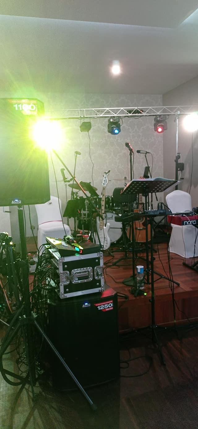 Scena z instrumentami zespołu muzycznego, w tym perkusja, gitara basowa, keyboard Nord, mikrofony na statywach, oświetlenie sceniczne i kolumna głośnikowa Alto, przygotowana do występu na weselu...