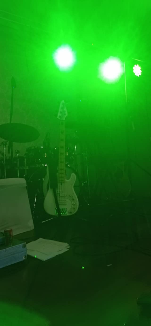 Biała gitara basowa na scenie, oświetlona intensywnym zielonym światłem, za nią widoczny zestaw perkusyjny.