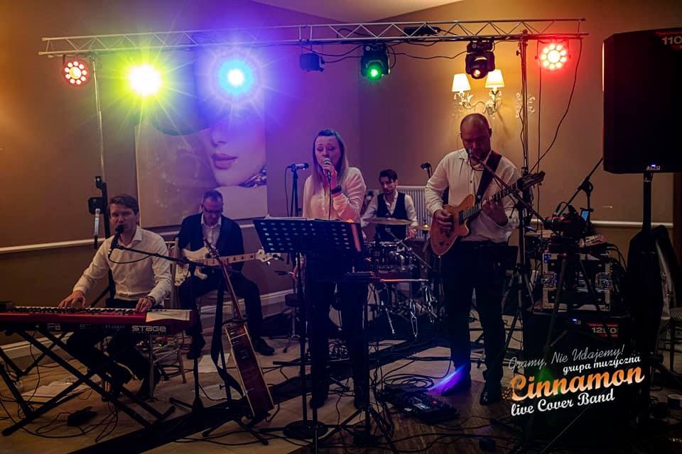 Zespół muzyczny w trakcie występu: wokalistka śpiewa do mikrofonu, gitarzysta gra na gitarze elektrycznej, klawiszowiec gra na keyboardzie, a perkusista na perkusji. Scena oświetlona kolorowymi...