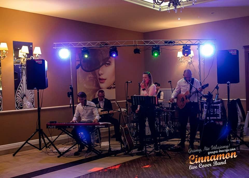 Zespół muzyczny grający na żywo na scenie podczas wydarzenia, widoczni muzycy grający na gitarze, keyboardzie, perkusji oraz wokalistka z mikrofonem, oświetlenie sceniczne, w tle dekoracja ze...