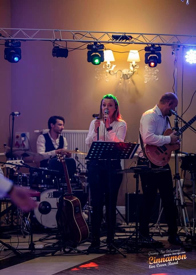 Zespół muzyczny w trakcie występu na scenie: wokalistka śpiewa do mikrofonu, gitarzysta gra na gitarze elektrycznej, a perkusista gra na perkusji. Scena oświetlona reflektorami.