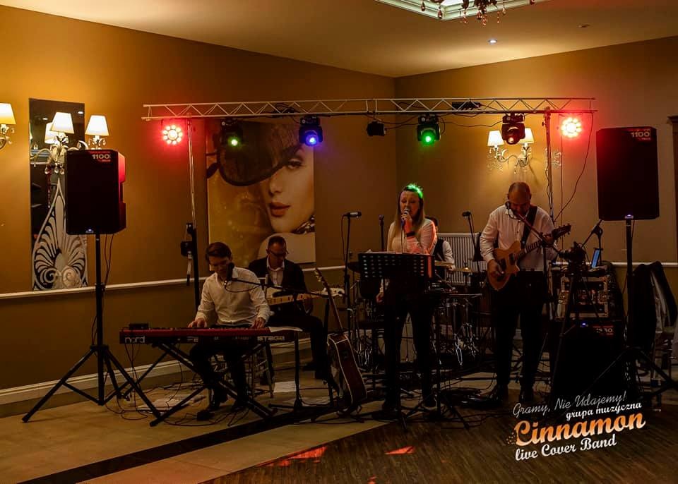 Zespół muzyczny Cinnamon Live Cover Band w trakcie występu na sali bankietowej, oświetlenie sceniczne, wokalistka z mikrofonem, klawiszowiec gra na Nord Stage, gitarzysta z gitarą akustyczną...