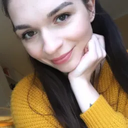 Portret uśmiechniętej brunetki w musztardowym swetrze, opierającej brodę na dłoni, z delikatnym tatuażem na nadgarstku.