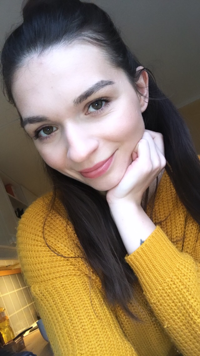 Portret uśmiechniętej brunetki w musztardowym swetrze, opierającej brodę na dłoni, z delikatnym tatuażem na nadgarstku.