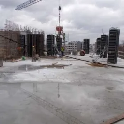 Wylewka betonowa na budowie, widoczne szalunki, zbrojenia i dźwig z pojemnikiem na beton, odbicie żurawia w mokrej powierzchni betonu, pracownicy w żółtych kamizelkach.