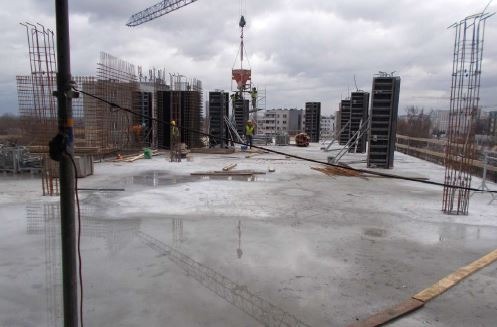 Wylewka betonowa na budowie, widoczne szalunki, zbrojenia i dźwig z pojemnikiem na beton, odbicie żurawia w mokrej powierzchni betonu, pracownicy w żółtych kamizelkach.