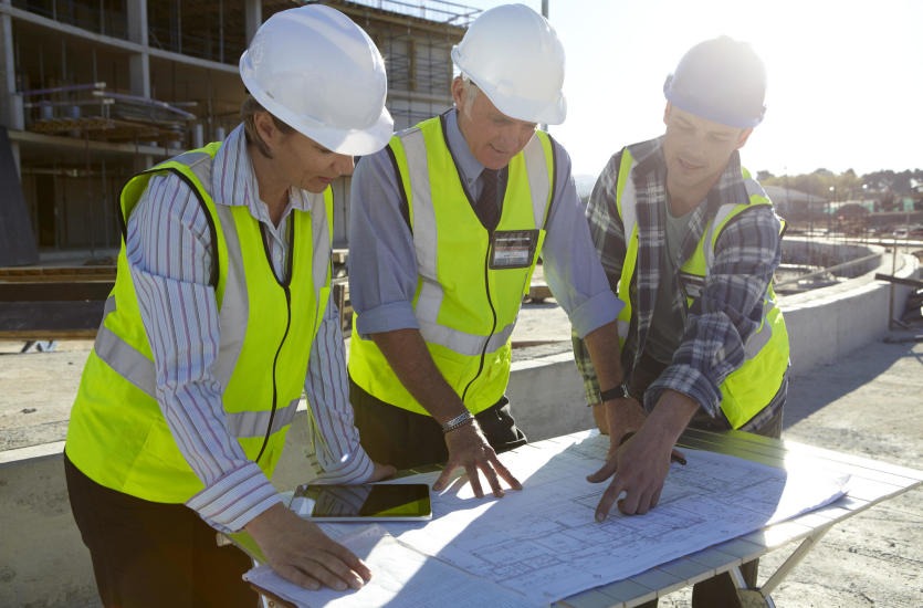Trzech inżynierów budowlanych w kaskach i kamizelkach odblaskowych analizuje plan budowy na placu budowy, z widocznym szkieletem budynku w tle.