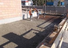 Prace budowlane na placu: dwóch robotników w pomarańczowych kamizelkach rozprowadza beton na siatce zbrojeniowej, widoczne drewniane szalunki i ceglany fragment budynku w tle.