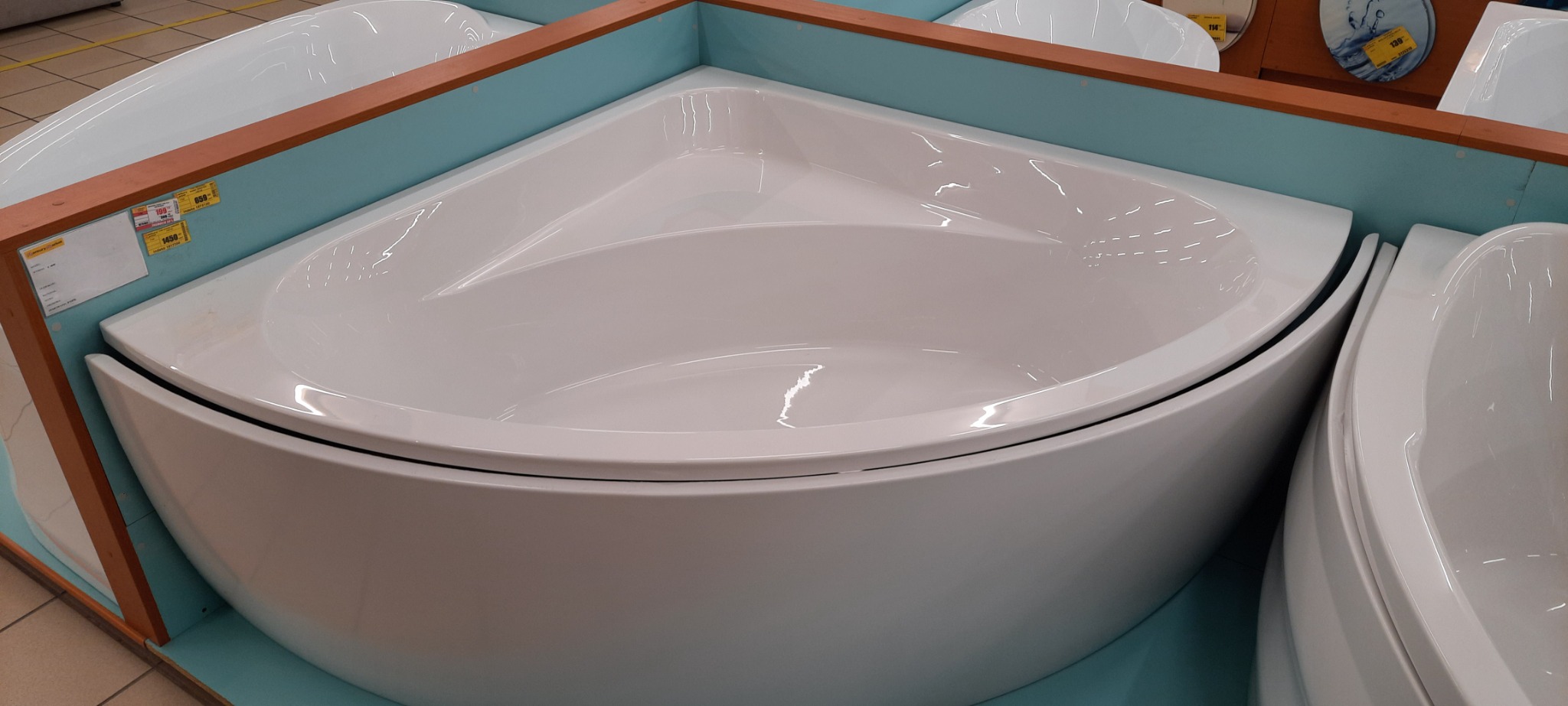 Biała, narożna wanna akrylowa, ustawiona na ekspozycji w sklepie, otoczona drewnianą i turkusową obudową.
