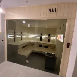 Nowoczesna sauna z przeszklonymi drzwiami przesuwnymi, drewnianymi ławkami w kształcie litery L i piecem elektrycznym z kamieniami, widoczny panel sterowania temperaturą.