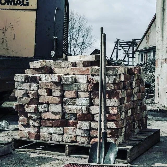 Stos cegieł ułożonych na palecie z dwiema łopatami opartymi o stos, w tle częściowo rozebrany budynek z widoczną konstrukcją dachu.