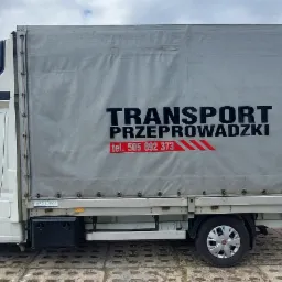 Biały samoch&oacute;d dostawczy z plandeką, na kt&oacute;rej widnieje napis 'TRANSPORT PRZEPROWADZKI' oraz numer telefonu, zaparkowany na betonowej nawierzchni, z zielenią w tle.