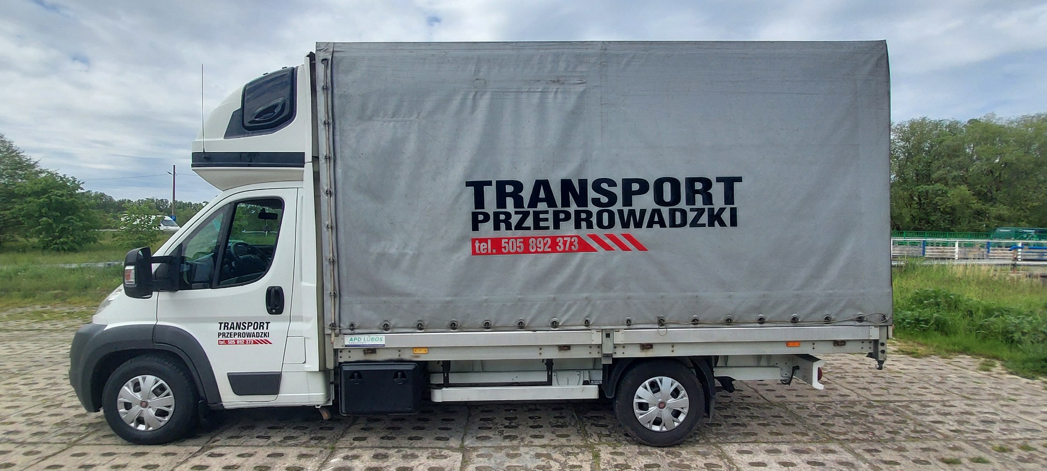 Biały samochód dostawczy z plandeką, na której widnieje napis 'TRANSPORT PRZEPROWADZKI' oraz numer telefonu, zaparkowany na betonowej nawierzchni, z zielenią w tle.