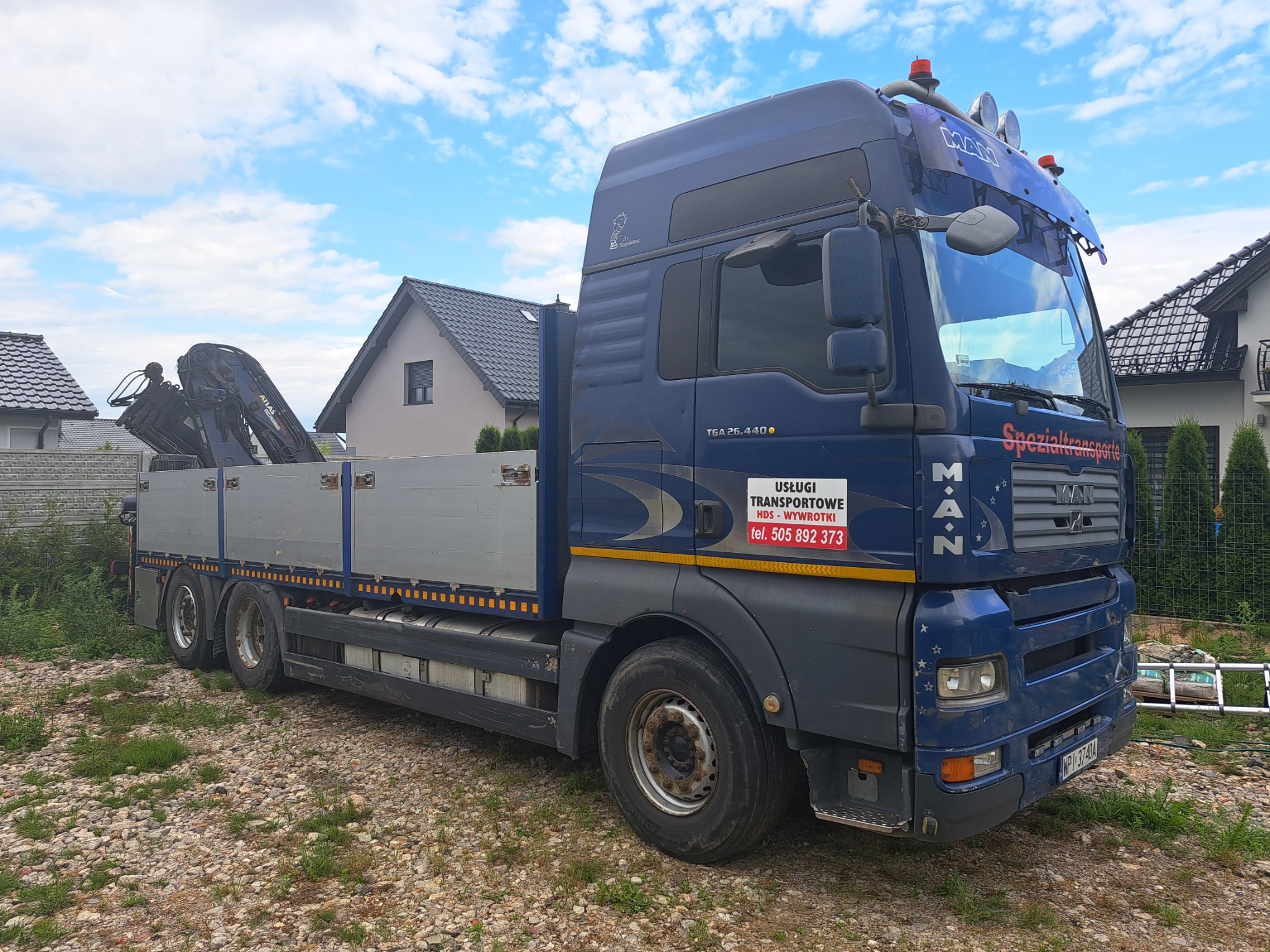Niebieska ciężarówka MAN z dźwigiem HDS na platformie, zaparkowana na żwirowym podłożu, w tle domy jednorodzinne i błękitne niebo z chmurami.