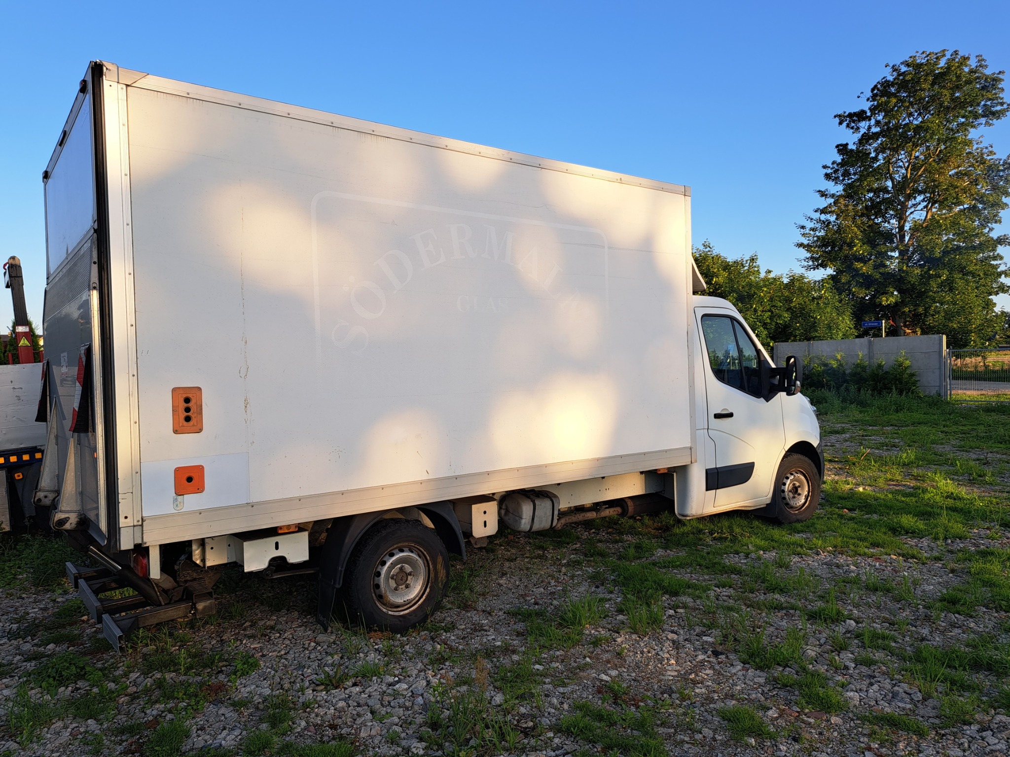 Biały samochód dostawczy z nadrukowanym, lekko rozmytym logo firmy 'Södermalm Glas' na boku, zaparkowany na trawiastym terenie w słoneczny dzień.