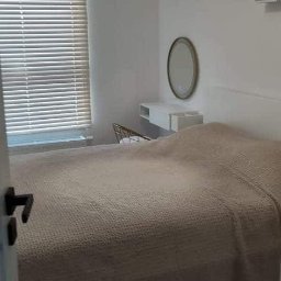 Artur Godlewski Usługi Remontowo-Budowlane - Minimalistyczna sypialnia w bieli z drewnianą podłogą i elementami dekoracyjnymi: lampa z abażurem z naturalnego materiału, lustro nad toaletką, półki ścienne. Widok zza uchylonych drzwi.