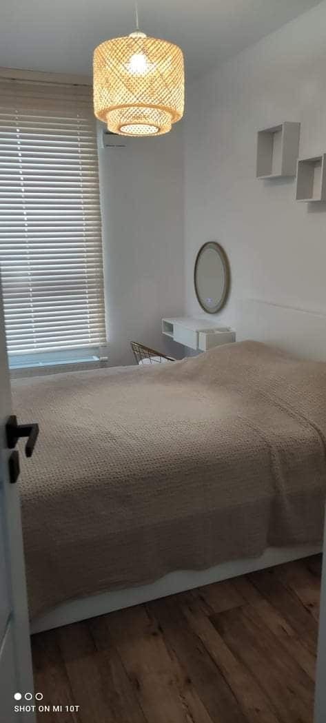 Minimalistyczna sypialnia w bieli z drewnianą podłogą i elementami dekoracyjnymi: lampa z abażurem z naturalnego materiału, lustro nad toaletką, półki ścienne. Widok zza uchylonych drzwi.