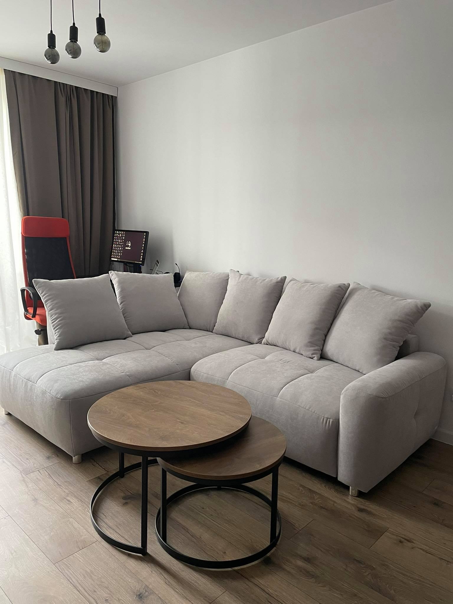 Jasnoszara sofa narożna z poduszkami i dwoma okrągłymi stolikami kawowymi w salonie z panelami podłogowymi i zasłonami w tle.