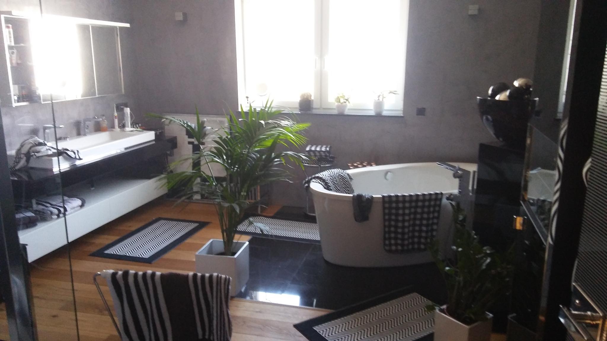 Nowoczesna łazienka z szarymi ścianami, wolnostojącą białą wanną, umywalką na czarnym blacie, drewnianą podłogą i czarno-białymi dywanikami oraz roślinami doniczkowymi.