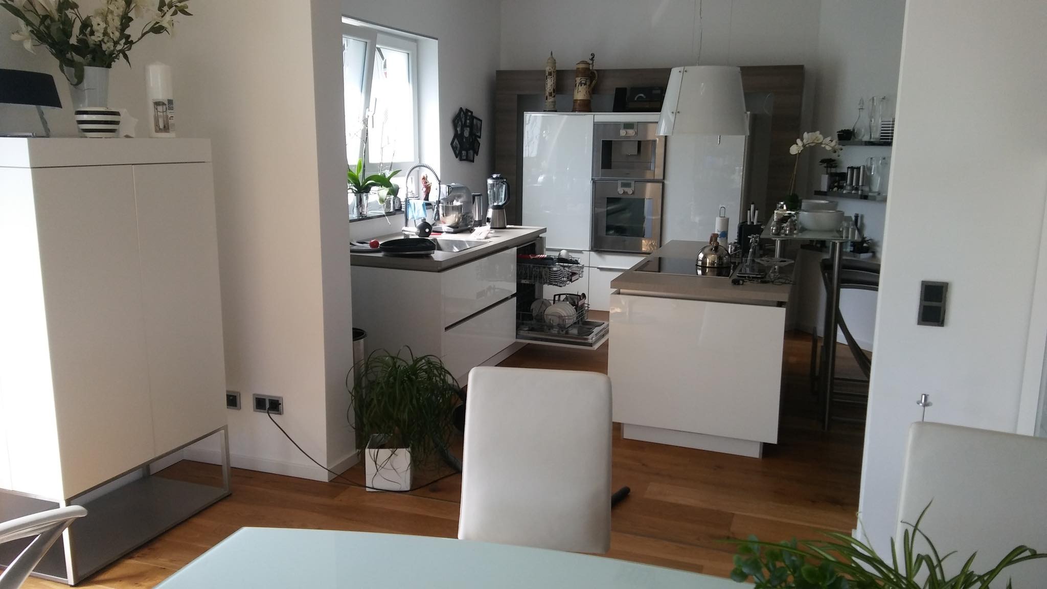 Jasna, nowoczesna kuchnia z białymi meblami, drewnianą podłogą i otwartą zmywarką, widok na okno z roślinami i dekoracje.