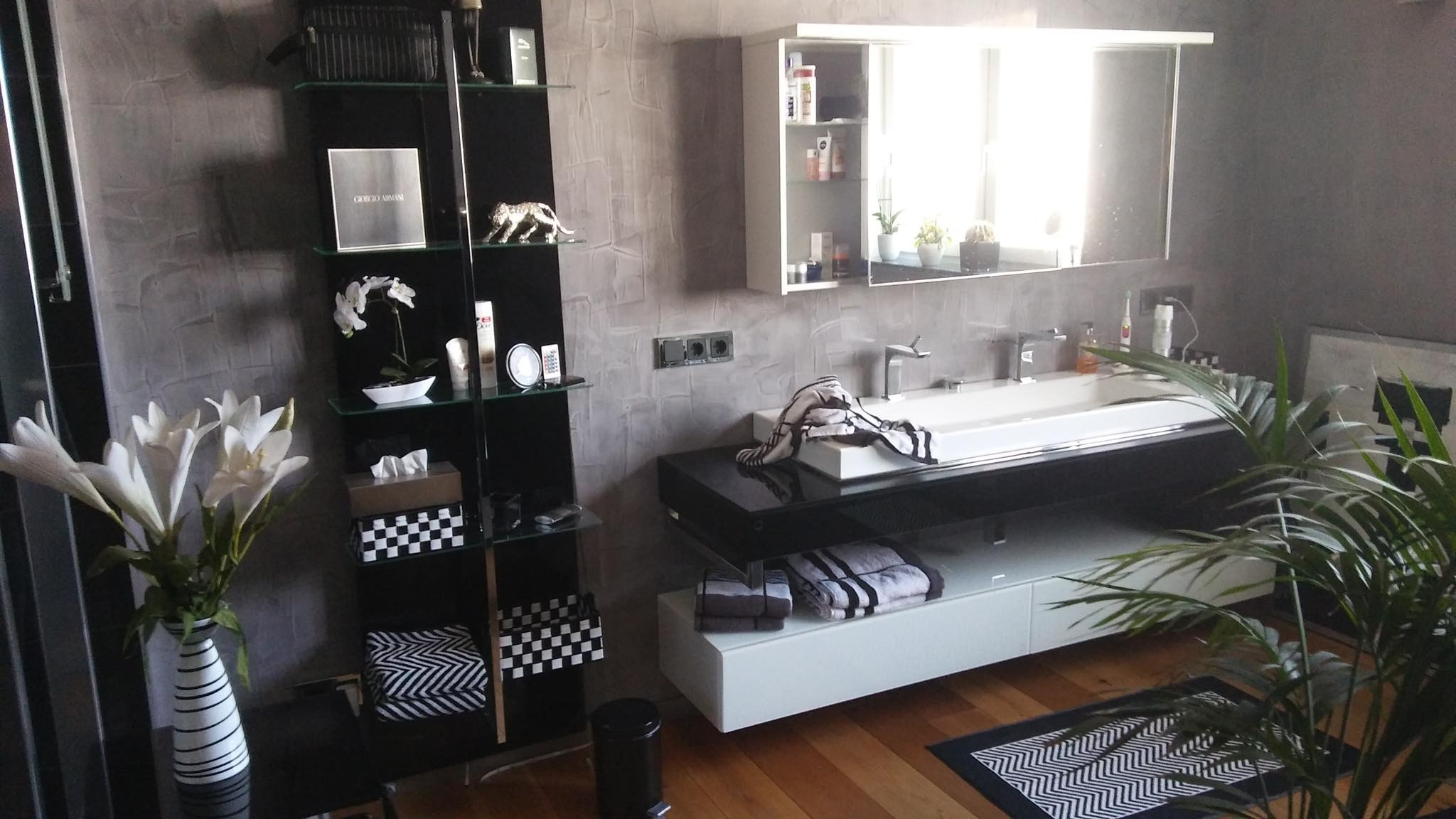 Nowoczesna łazienka z szarą ścianą o fakturze betonu, czarnym regałem z dekoracjami, białą umywalką z czarnym blatem i lustrem w szafce, uzupełniona o kwiaty w wazonie i doniczce.
