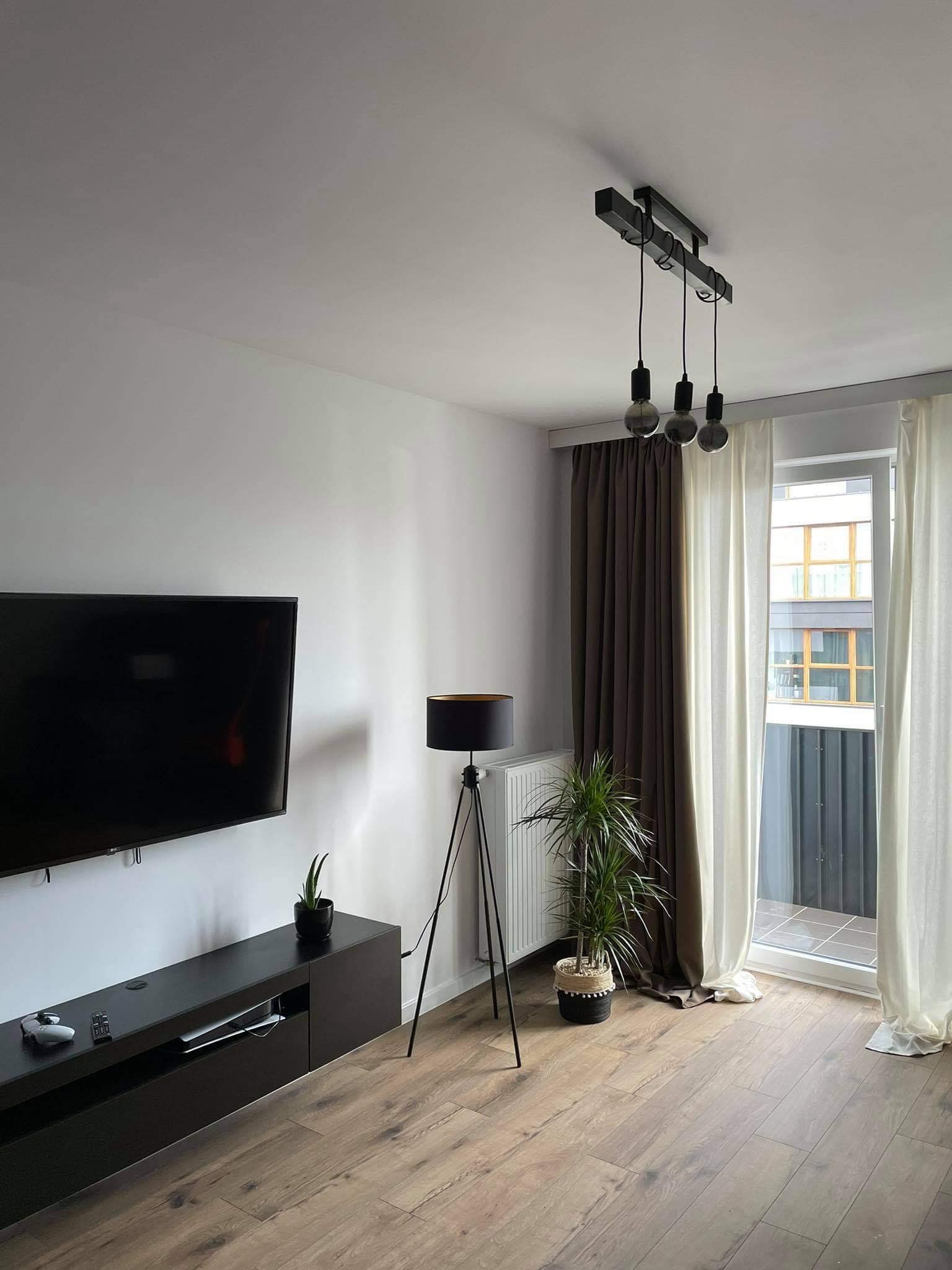 Nowoczesny salon z drewnianą podłogą, czarną szafką RTV, telewizorem, lampą trójnóg i roślinami doniczkowymi, okno z zasłonami w odcieniach beżu i brązu oraz potrójna lampa wisząca z żarówkami...